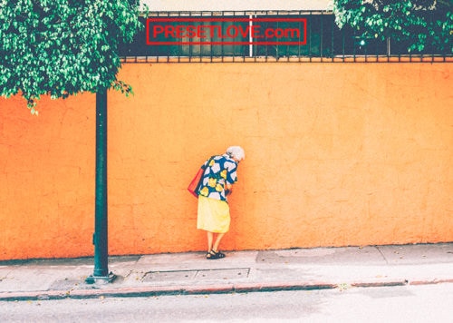 An old lady walking on the sidewalk beside a vibrant yellow wall